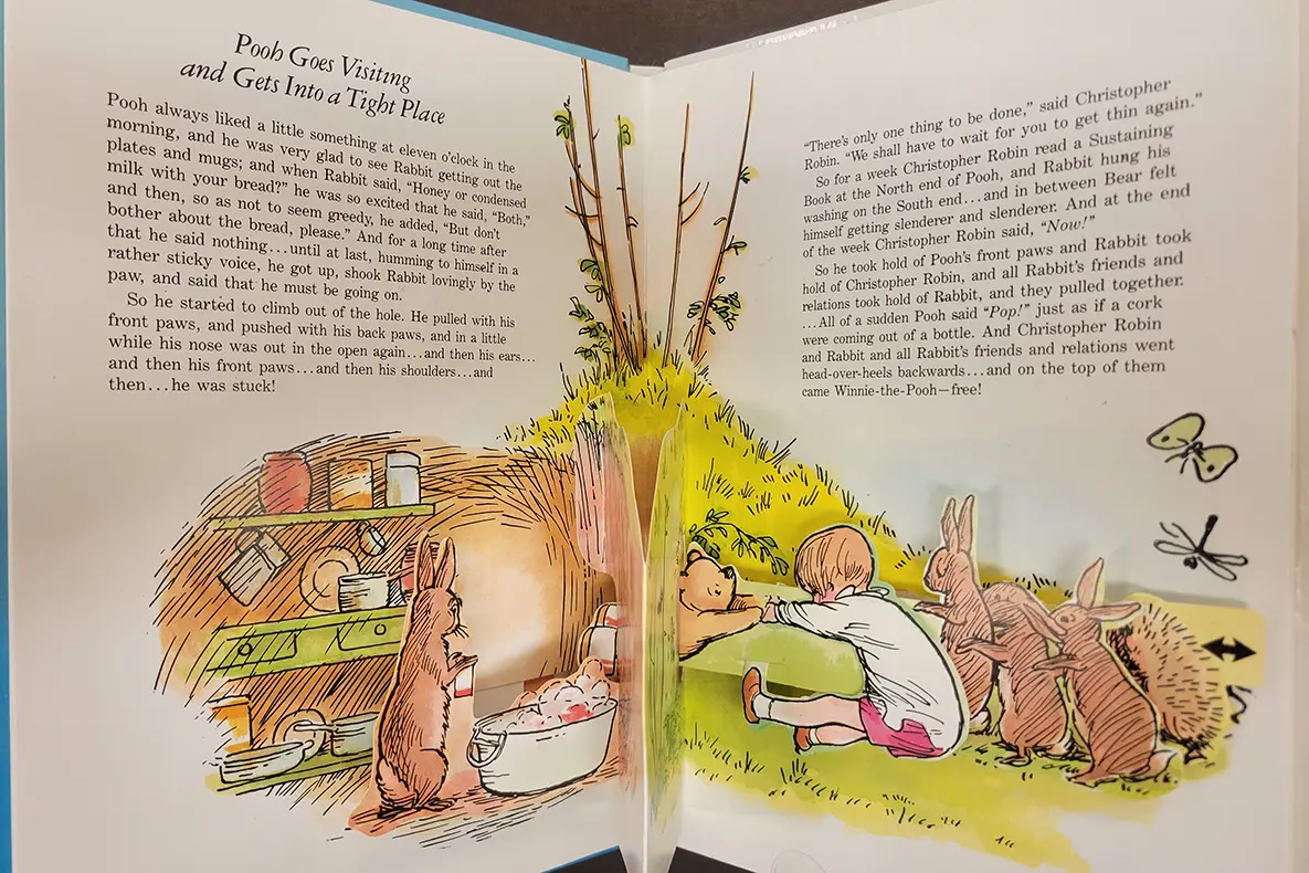 An open pop-up book that says, "Pooh Goes Visiting and Gets into a Tight Space" and an illustration with Pooh stuck in a hole with Christopher Robin trying to pull him out.
