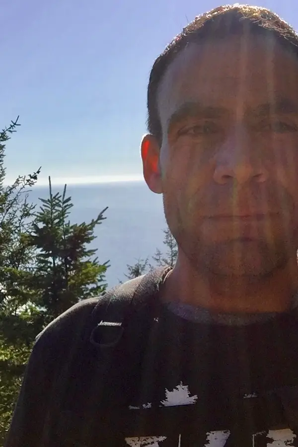 Picture of Craig from the shoulders up standing on a mountain in the Oregon Coast Range with the Pacific Ocean in the background and the sun shining overhead.