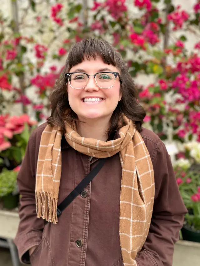 White woman with shoulder length brown hair and glasses wear a brown coat and tan scarf, standing against a background of red flowers.nt background. 