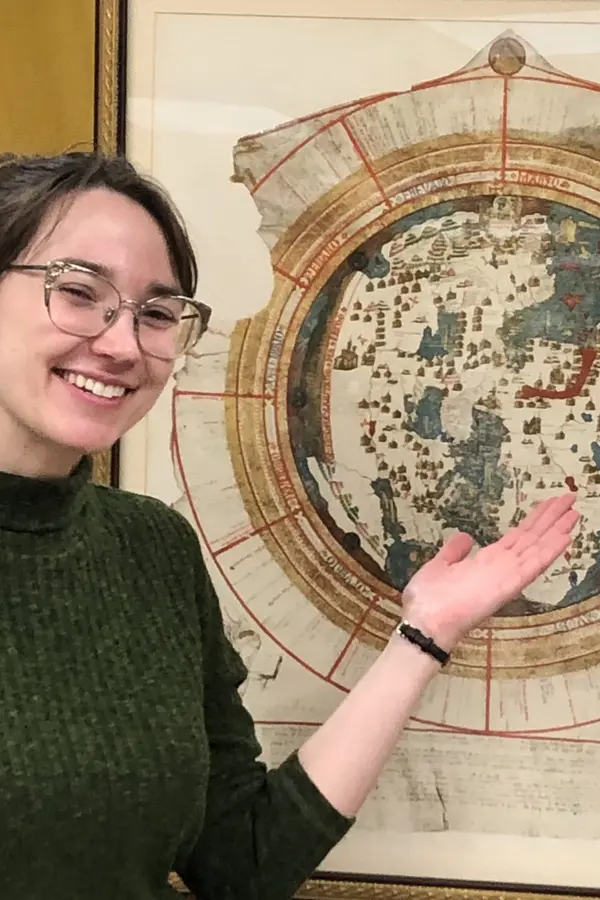 A white woman in a green turtleneck gestures behind her towards a circular map of the world from 1542.