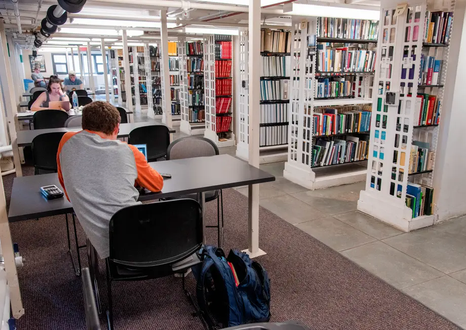 Students sitting at rectangular tables along a wall next to floor-to-ceiling book stacks.