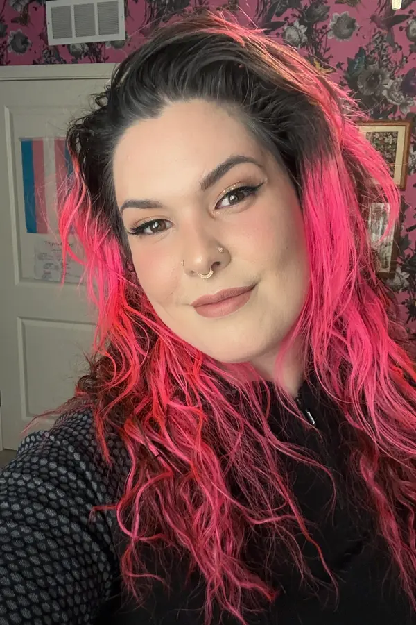 Smiling woman with light colored skin, peach colored lips, dark hair with bright pink highlights, and wearing a black shirt.
