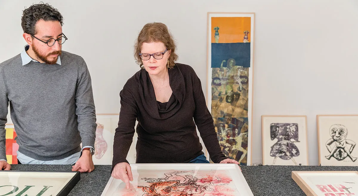 Woman gesturing to at a framed art piece to a man standing next to her.