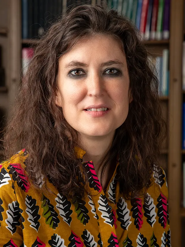 headshot of Evyn Kropf, a person with light skin, wavy dark hair, dark eyebrows and dark eyes, in front of a bookcase overflowing with books
