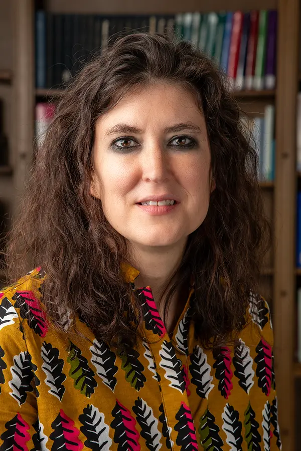 headshot of Evyn Kropf, a person with light skin, wavy dark hair, dark eyebrows and dark eyes, in front of a bookcase overflowing with books