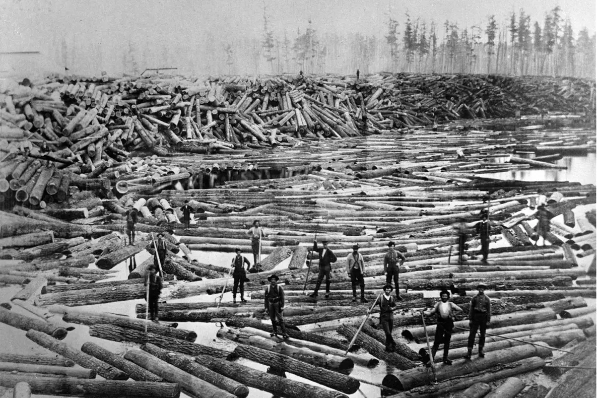 Men standing on logs that almost completely block a bay of water.