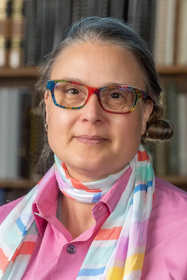 Photo of Marilène "Mars" De Ritis - a woman with greying hair and multi-colored eyeglass frames wearing a magenta shirt and light-weight scarf in front of Reference Reading Room bookshelf