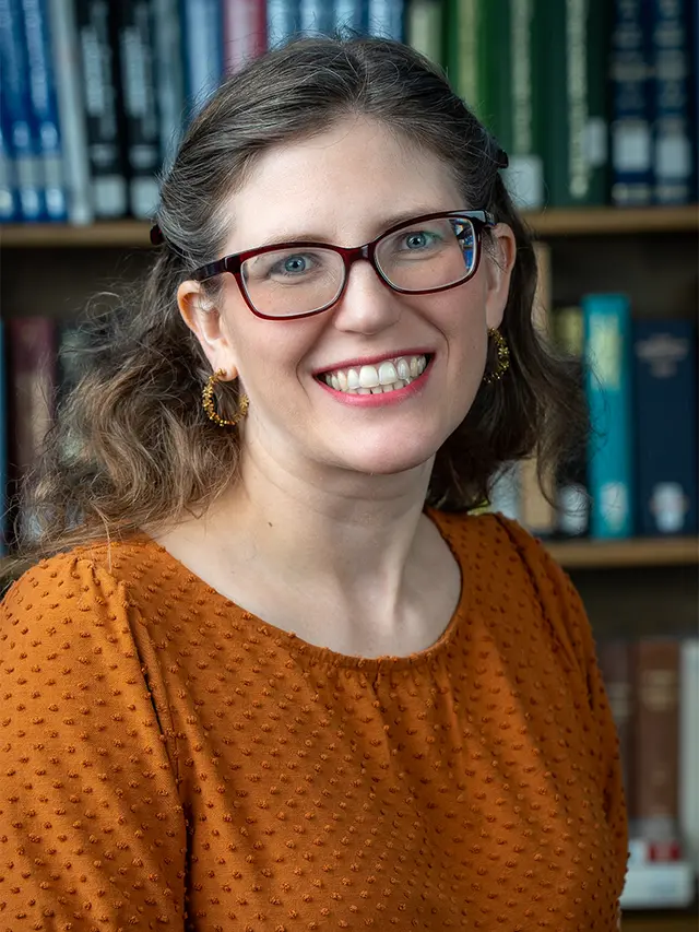 Photograph of Juli McLoone, a white woman with brown hair and glasses, wearing oranged blue eyes, wearing purple.