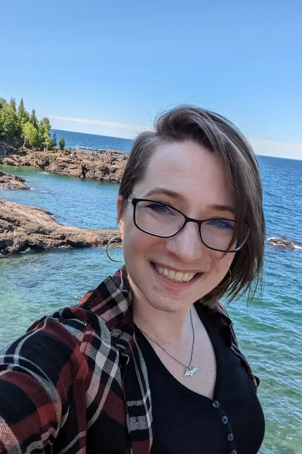 Photograph of Hilary Severyn, a white woman with short brown hair and glasses, in front of a rocky lakeshore.