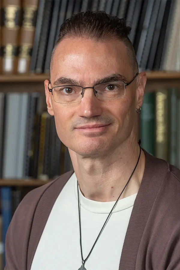 Photo of Scott Martin: a white man with glasses and short brown hair, wearing a white t-shirt and brown sweater, in front of a shelf of books