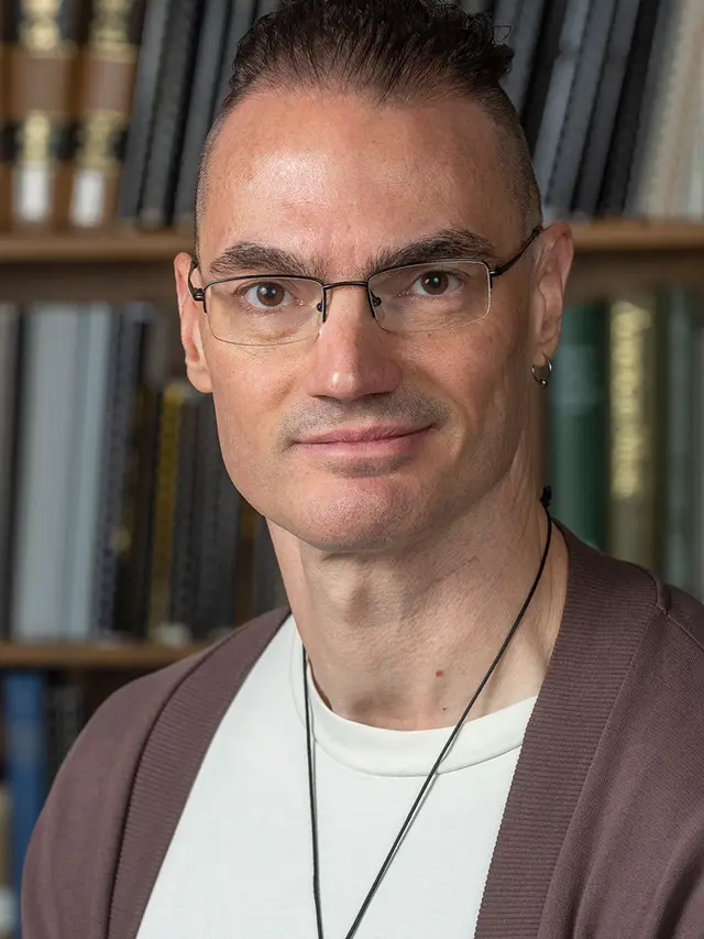 Photo of Scott Martin: a white man with glasses and short brown hair, wearing a white t-shirt and brown sweater, in front of a shelf of books