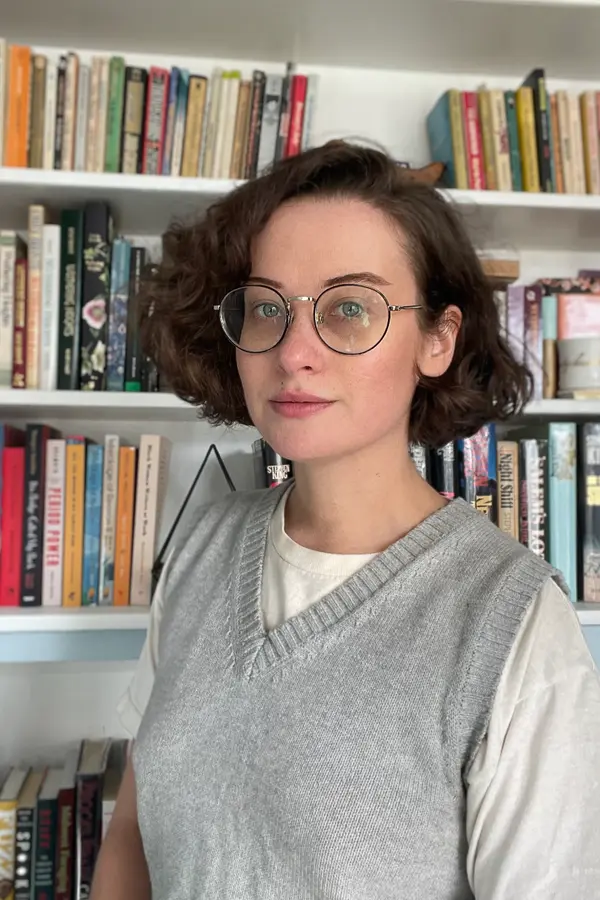 Marisa Mercurio standing in front of a bookshelf, wearing a grey sweater vest over a white t-shirt.