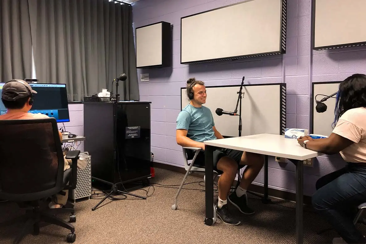 Students using the audio production equipment in the Perlstein Editing Room, with one a computer, and two talking into microphones with sound buffering panels behind them.