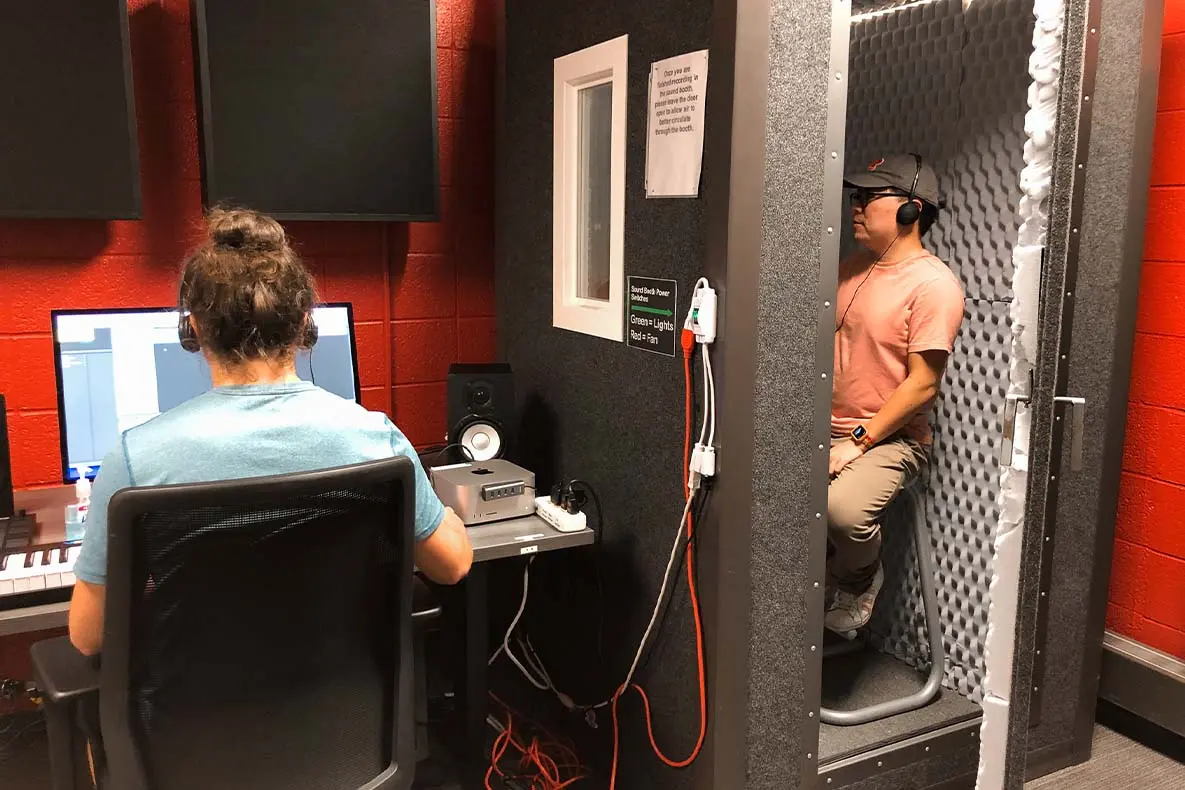 Students using the audio production equipment in the Winberg Room, with one in a sound booth and the other at a computer.