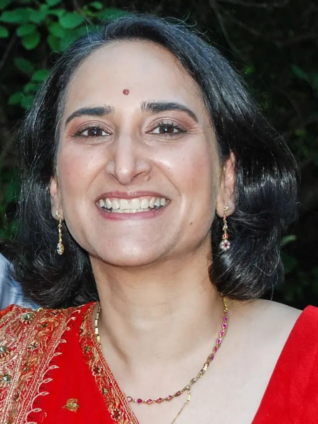 Head and shoulders image of a dark-haired woman with an olive complexion, wearing a red sari and smiling broadly