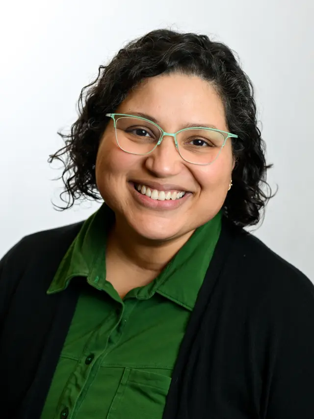 Photograph of Vitalina Robinson, a mixed-race latine woman with light brown skin, wearing glasses, a green dress, and smiling.