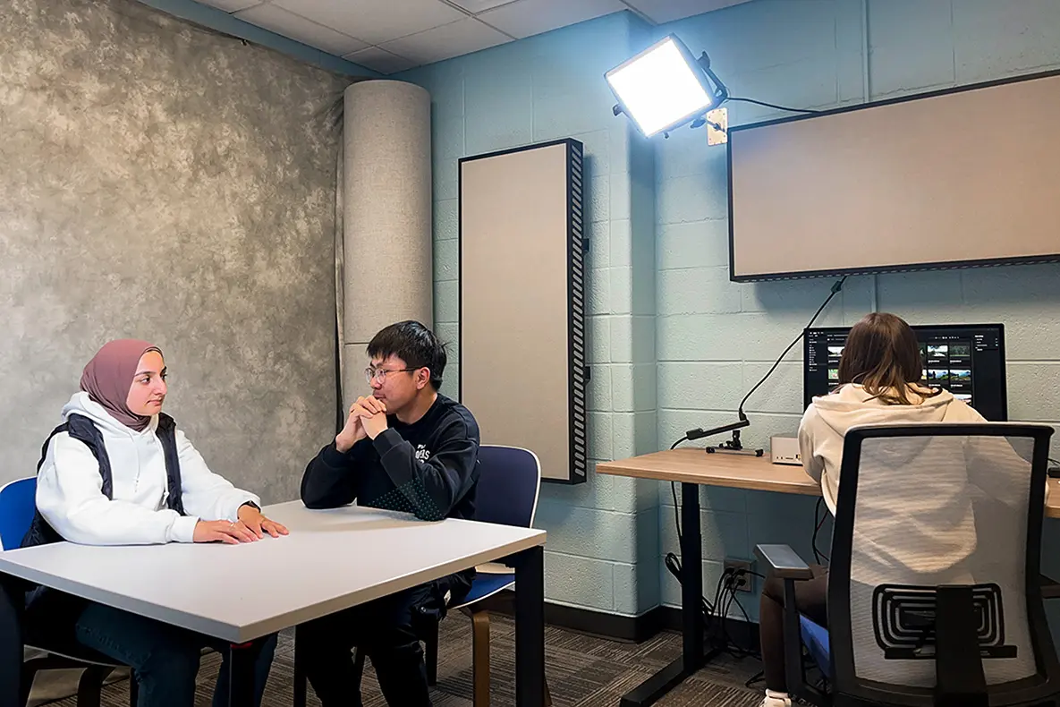 Two students talking at a table under a production light with another student working at a computer.