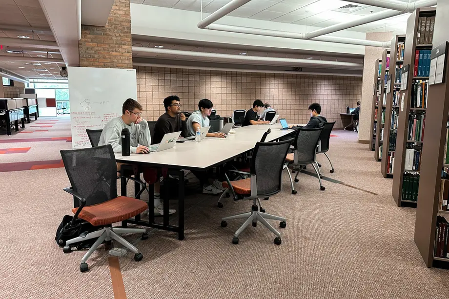 Students working at a long rectangular table next to book stacks with a whiteboard behind them and windows in the distance.