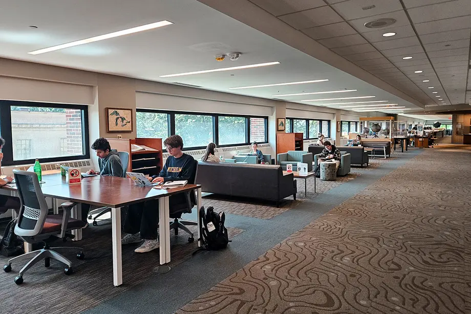 Students studying at rectangular tables in comfy chairs alongside windows.