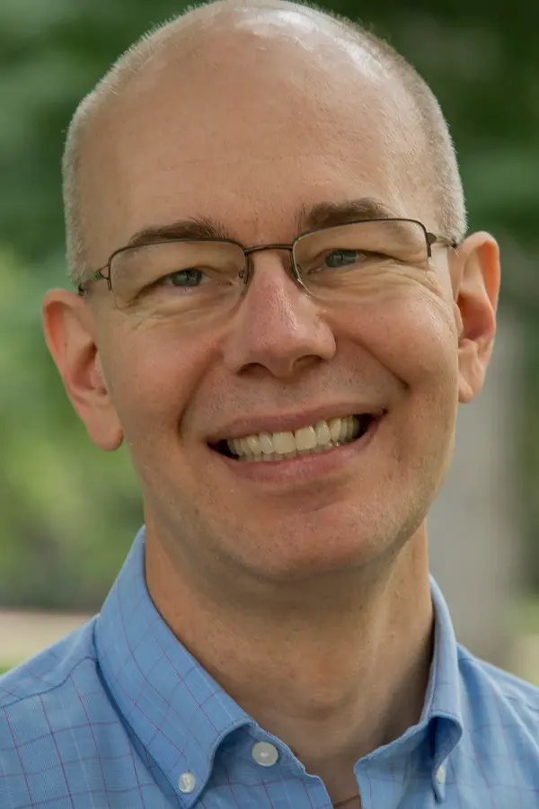 Photograph of Scott Dennis, a white man with a mostly bald head and very short gray hair on the sides, wearing clear glasses, a blue shirt, and a big smile in front of green foliage.