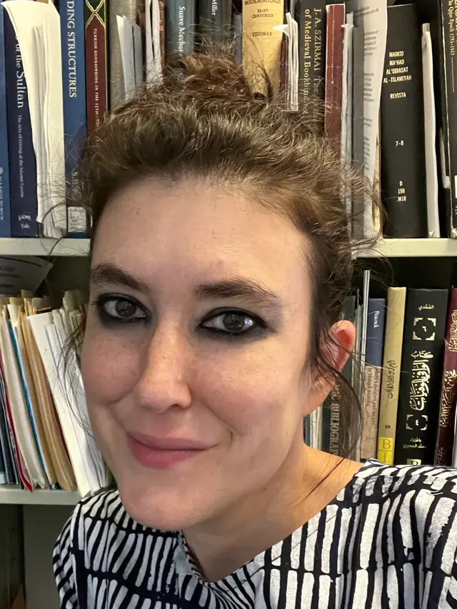 headshot of Evyn Kropf, a person with light skin, wavy dark hair, dark eyebrows and dark eyes, in front of a bookcase overflowing with books