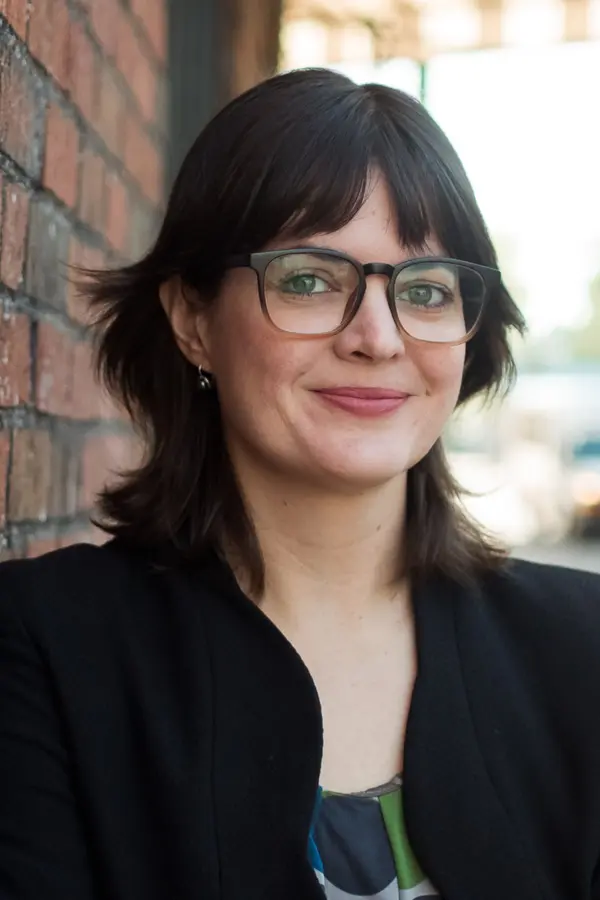 photo of smiling person with glasses and dark hair leaning against a brick wall