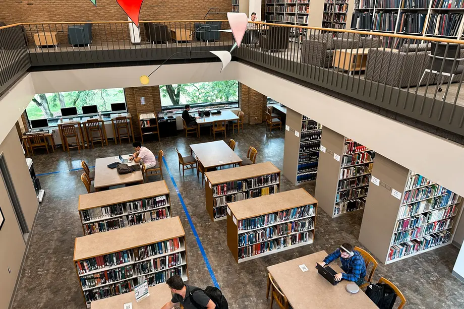 View from the top of two floors with an opening in the middle. The ground floor has rectangular tables and low bookshelves, the top floor has a railing and armchairs.