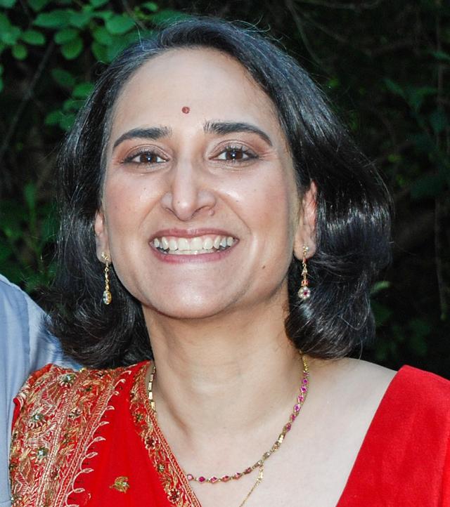 Head and shoulders image of a dark-haired woman with an olive complexion, wearing a red sari and smiling broadly