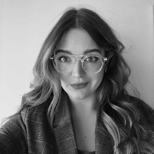 Black and white photo of a white woman with long hair, round clear glasses, and a checkered blazer.