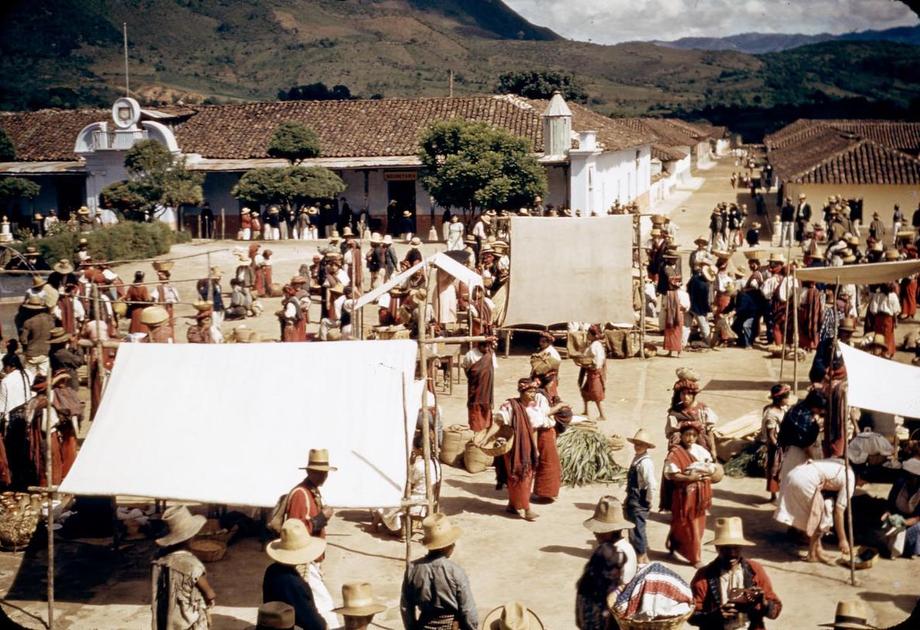 Market square with people amid a few tented stalls, white buildings in the background, and mountains in the distance