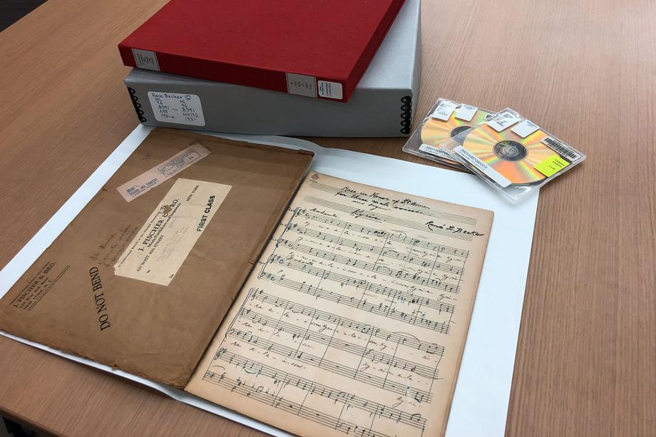 Sheet music with annotations and an original envelope, a stack of archival boxes, and CDs from the Voice of America collection.