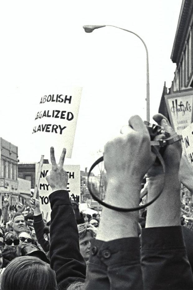 Black and white photograph from within a crowd of protestors with many raised arms visible. A sign with "Abolish legalized slavery" is in the distance.