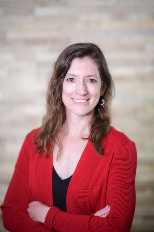 Photo of Emily Lynema, a white woman with long wavy brown hair and brown eyes wearing a red sweater