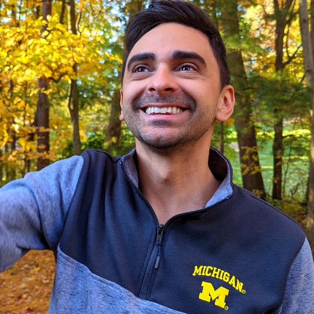 Josh wearing a University of Michigan sweater and smiling while standing outdoors with fall-colored trees in the background