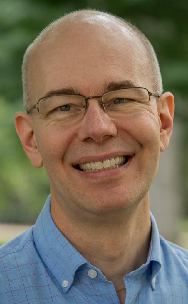 Photograph of Scott Dennis, a white man with a mostly bald head and very short gray hair on the sides, wearing clear glasses, a blue shirt, and a big smile in front of green foliage.