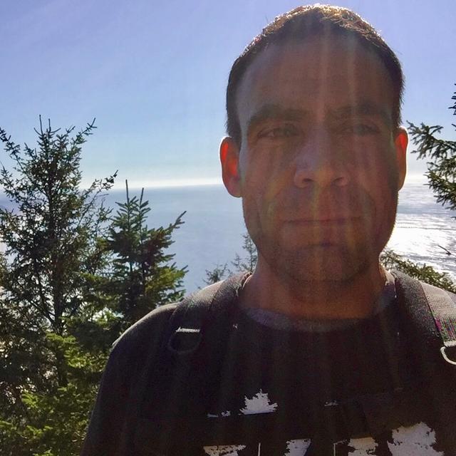 Picture of Craig from the shoulders up standing on a mountain in the Oregon Coast Range with the Pacific Ocean in the background and the sun shining overhead.