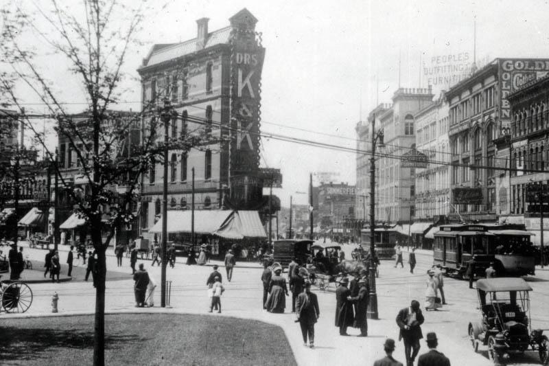 Black and white photograph from the early 1900s of a pedestrian intersection with people walking and buildings around it.