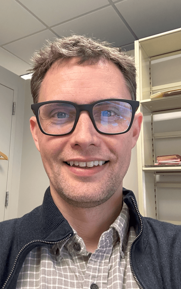 A smiling, bespectacled man in front of a bookcase