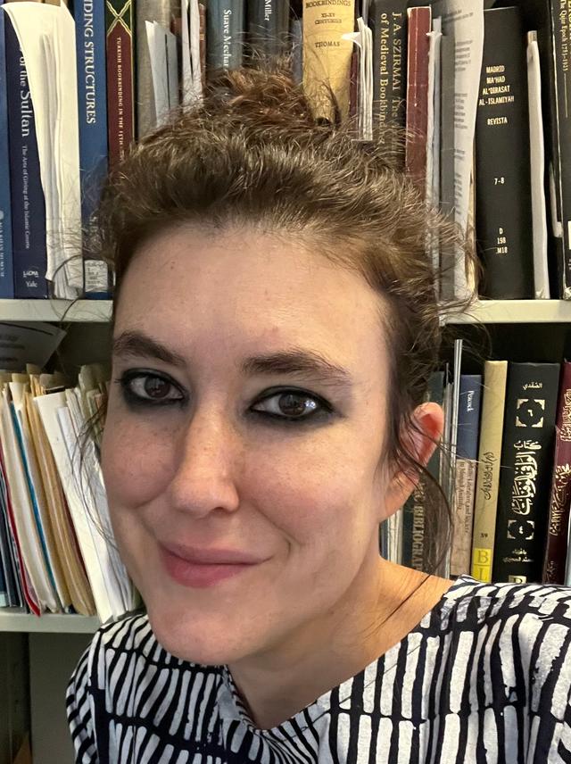 headshot of Evyn Kropf, a person with light skin, wavy dark hair, dark eyebrows and dark eyes, in front of a bookcase overflowing with books