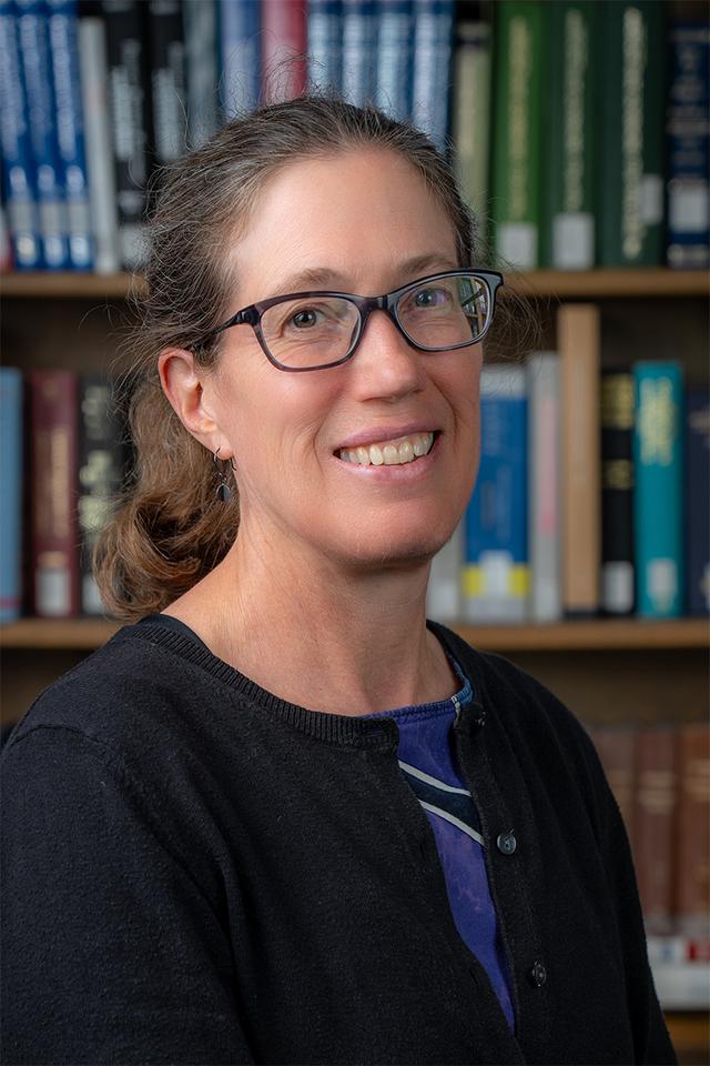 A photo of a white woman with a ponytail and glasses, wearing a multicolored dress and a black sweater.