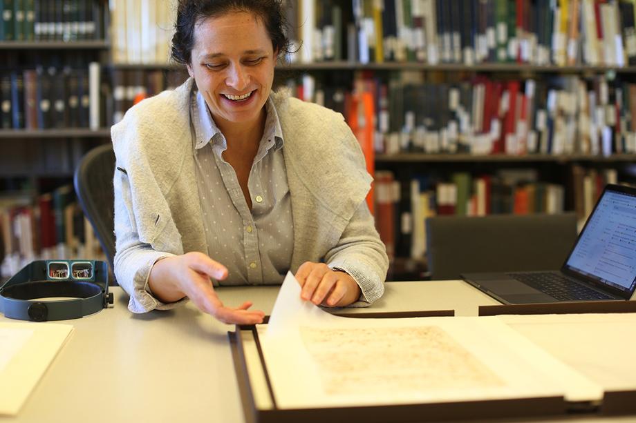 Smiling woman sitting at a desk and lifting the edge of a large printed sheet of paper from a manuscript box.