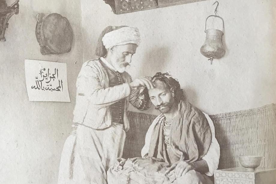 Gelatin silver photo print of a barber standing over a seated man, cutting his hair.