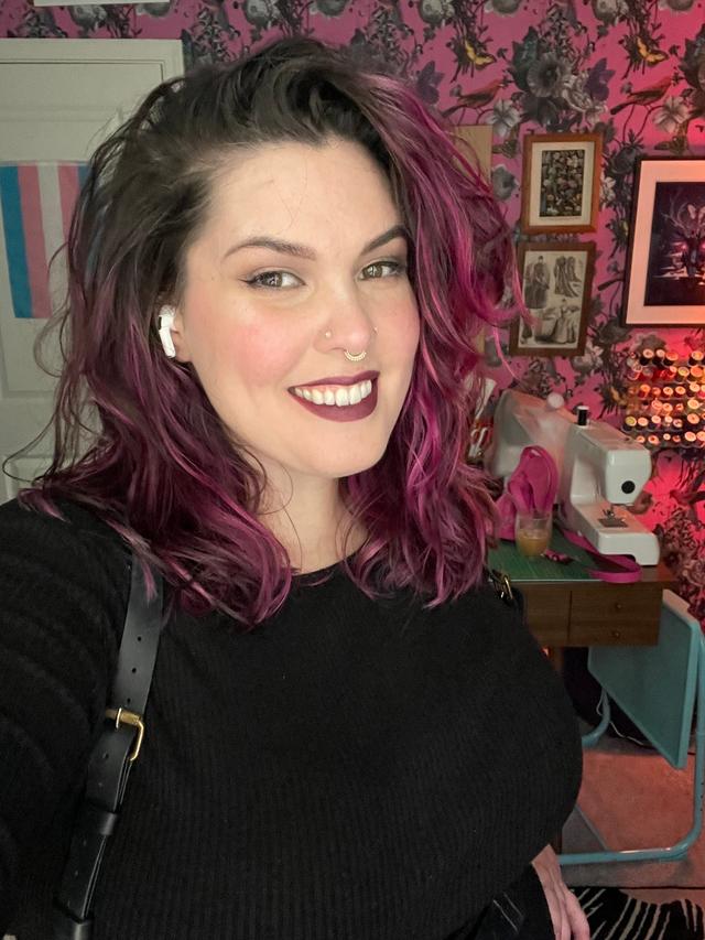 Smiling woman with light colored skin, dark purple lips, dark hair with pink highlights, and wearing a black shirt.