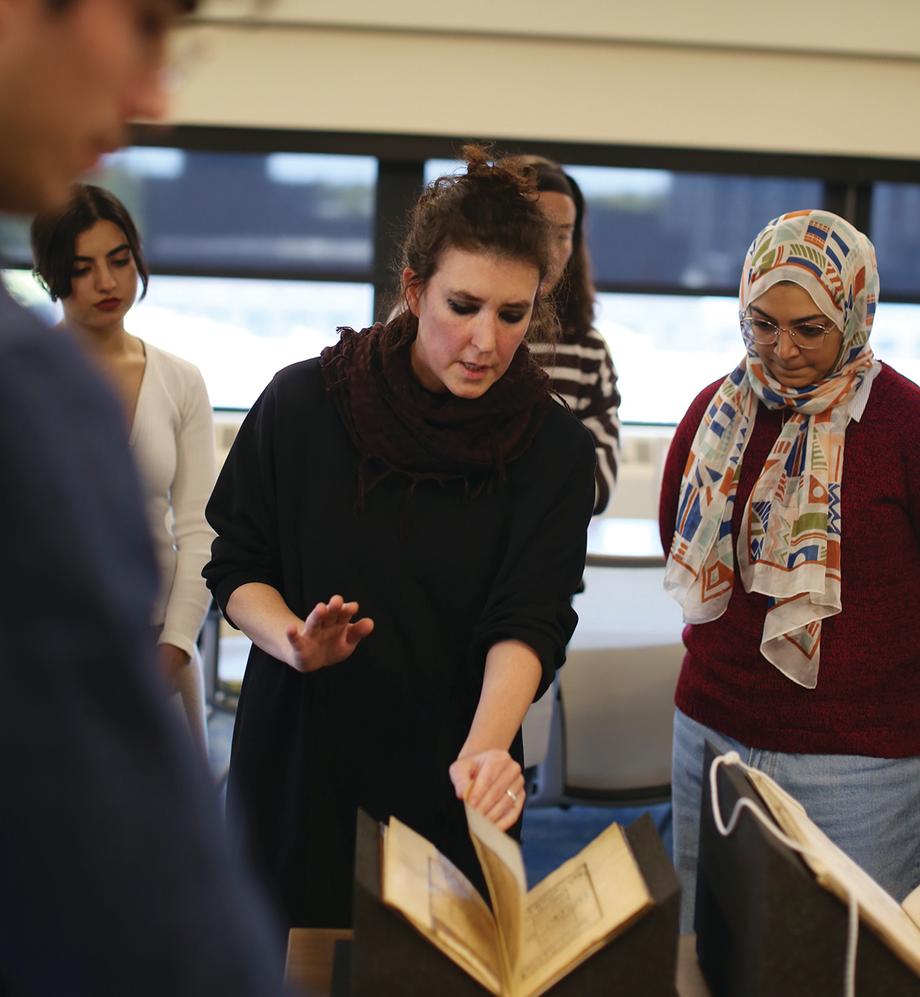 Woman turns the pages of a volume of Islamic manuscripts, while four of her students look on.