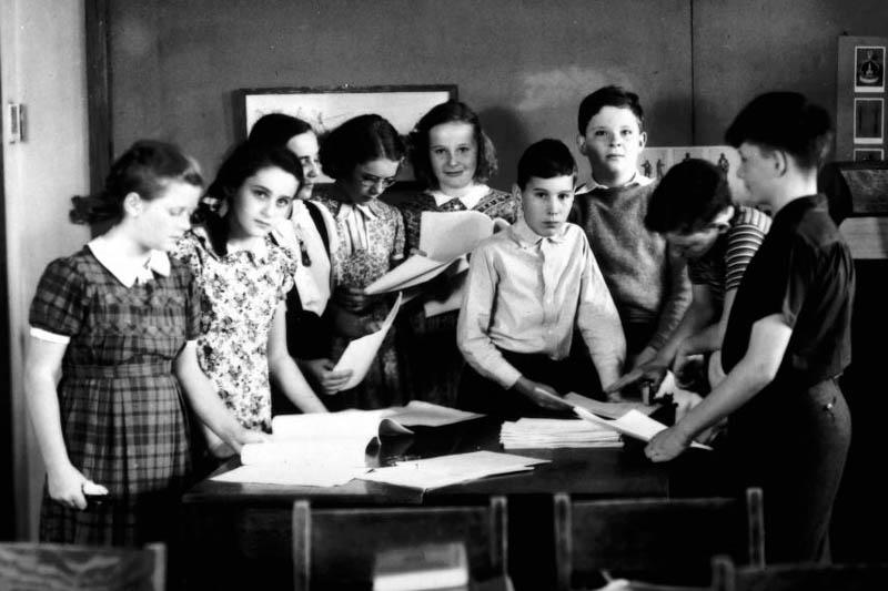 Black and white photo of a teacher and students gathered around a table looking at a lot of papers.