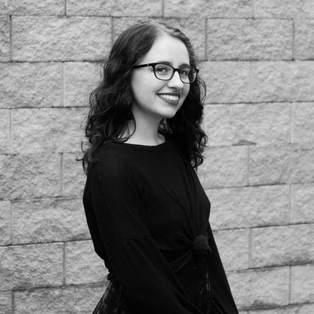 Grayscale photograph of Haley Winkle, a young white woman, smiling and wearing glasses along with a black shirt. She is standing against a concrete brick wall.