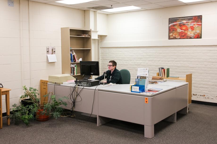 Staff member sitting behind a computer at a small desk in the Buhr Reading Room where there is art on the wall and plants on the floor.