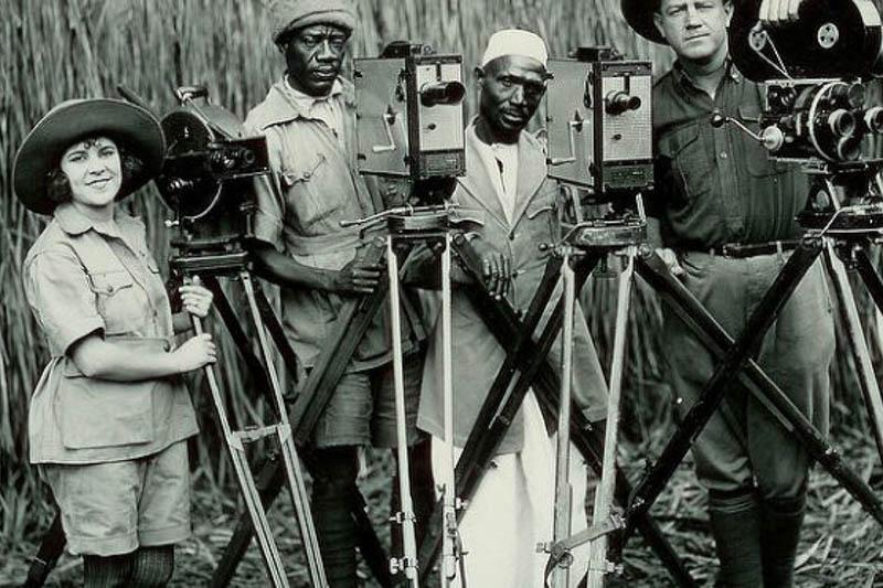 Osa and Martin Johnson, with two other men, all standing behind cameras with tall grasses in the background.