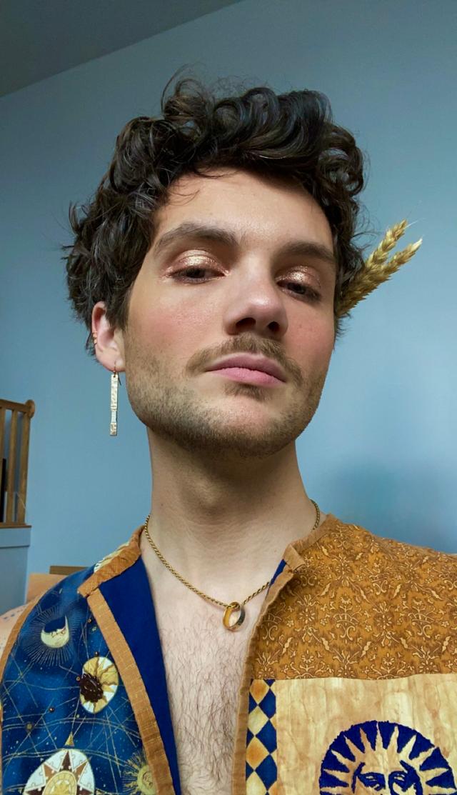 Close-up of j. Oceano — with short, dark curly hair and light facial hair, wearing shimmery gold eye makeup. They look slightly down at the camera with a neutral expression, wearing one long gold earring, a thin gold chain necklace with a small ring pendant, and a patterned vest in blue and gold with sun-and-moon motifs. A sprig of dried wheat is tucked behind their left ear; a pale blue wall and a wooden stair rail are in the background.