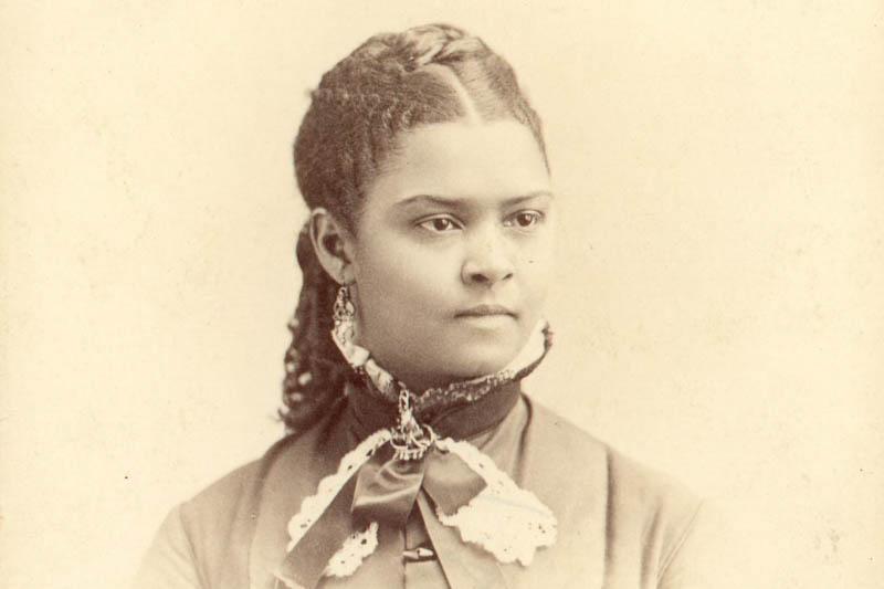 Monochrome seated portrait photograph of an African American woman with braids in her hair and high collared dress.  